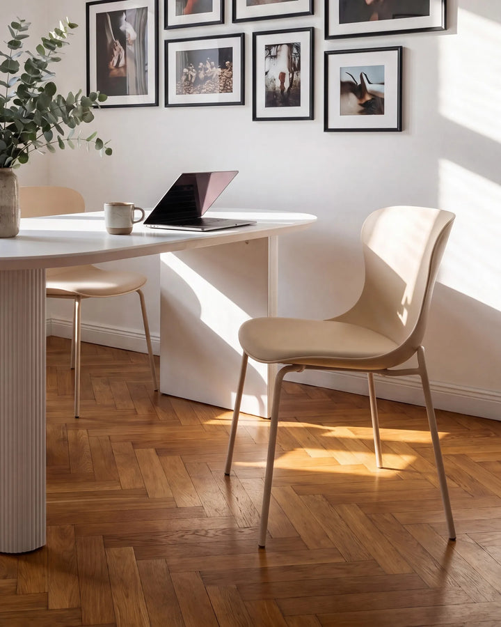 A modern beige upholstered dining chair with slim metal legs sits beside a white table in a sunlit room with wood herringbone flooring and framed wall art.