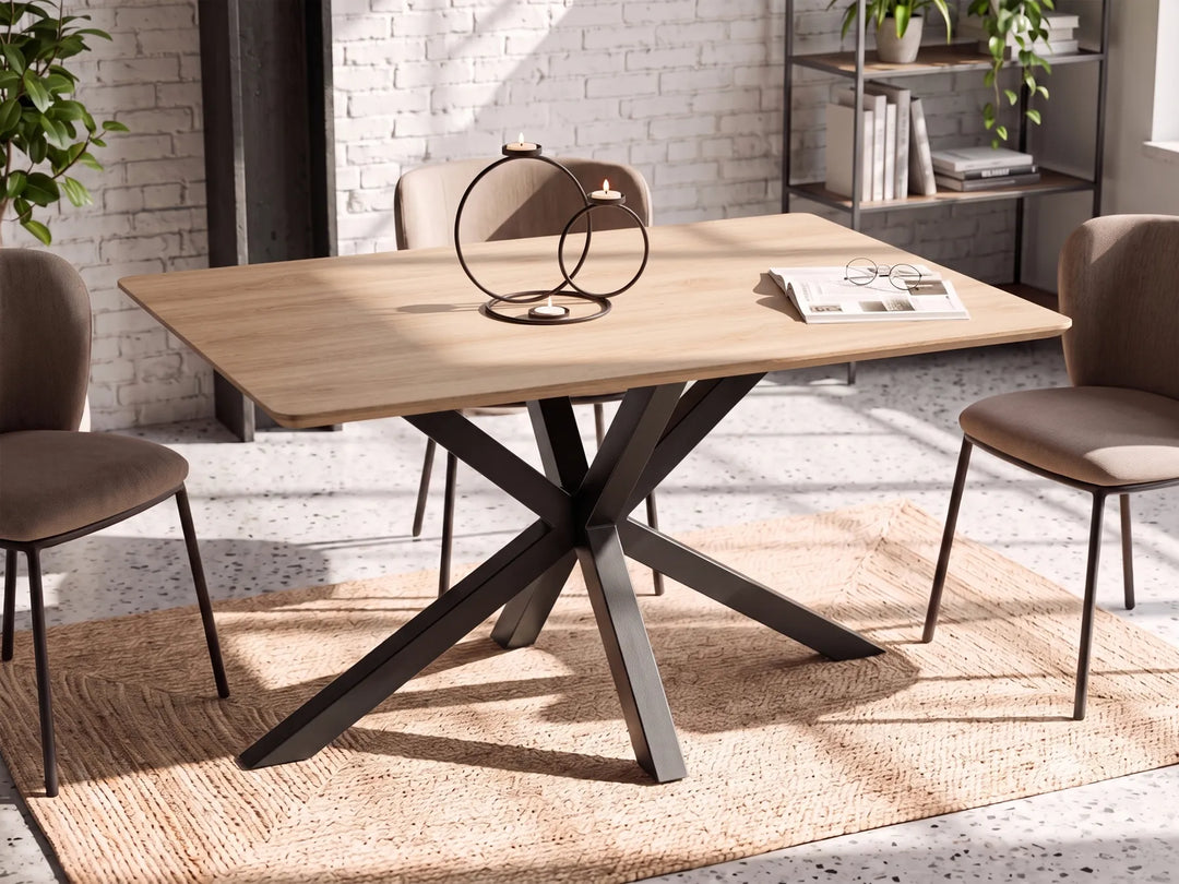 A rectangular dining table with a light natural wood top and a dark metal X-shaped pedestal base, surrounded by four upholstered dining chairs in a bright, modern room with a white brick wall and a woven rug.