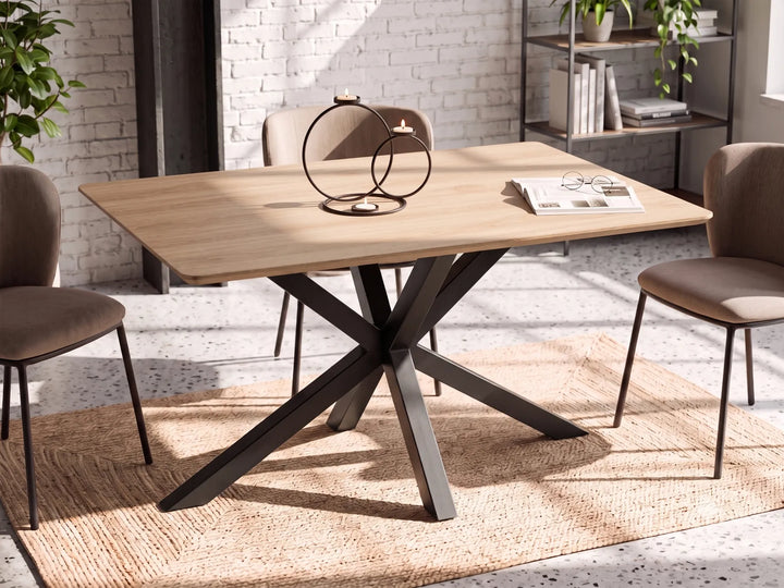 A rectangular dining table with a light natural wood top and a dark metal X-shaped pedestal base, surrounded by four upholstered dining chairs in a bright, modern room with a white brick wall and a woven rug.