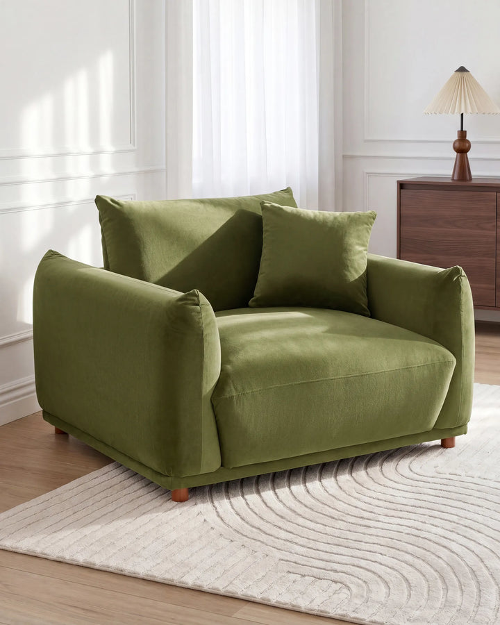 An olive green velvet armchair with two matching throw pillows, styled in a bright, modern room on a textured cream rug.
