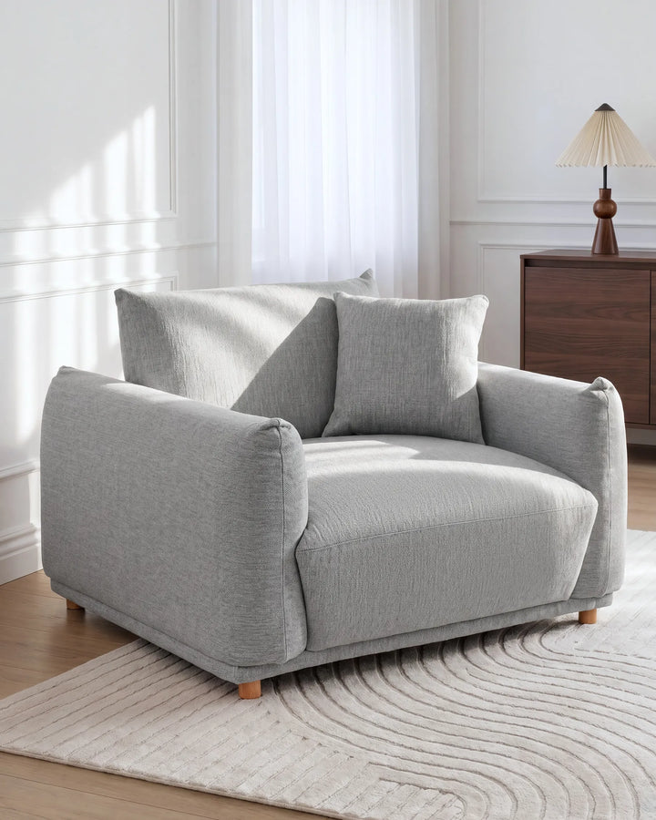 A light gray upholstered armchair with two matching back cushions, shown in a bright living room setting on a textured rug beside a wooden cabinet and lamp.
