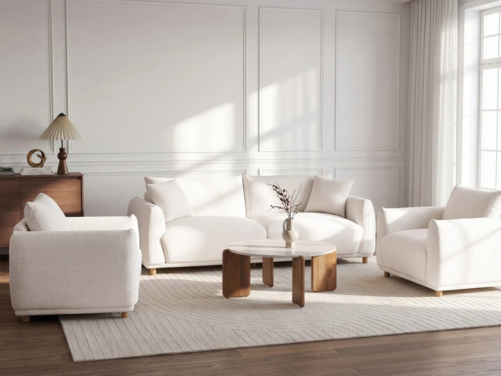 A bright, modern living room featuring a white three-seater sofa, a matching armchair, and a wooden coffee table on a light beige rug. The room has white paneled walls, large windows with sheer curtains, and warm natural light streaming in.