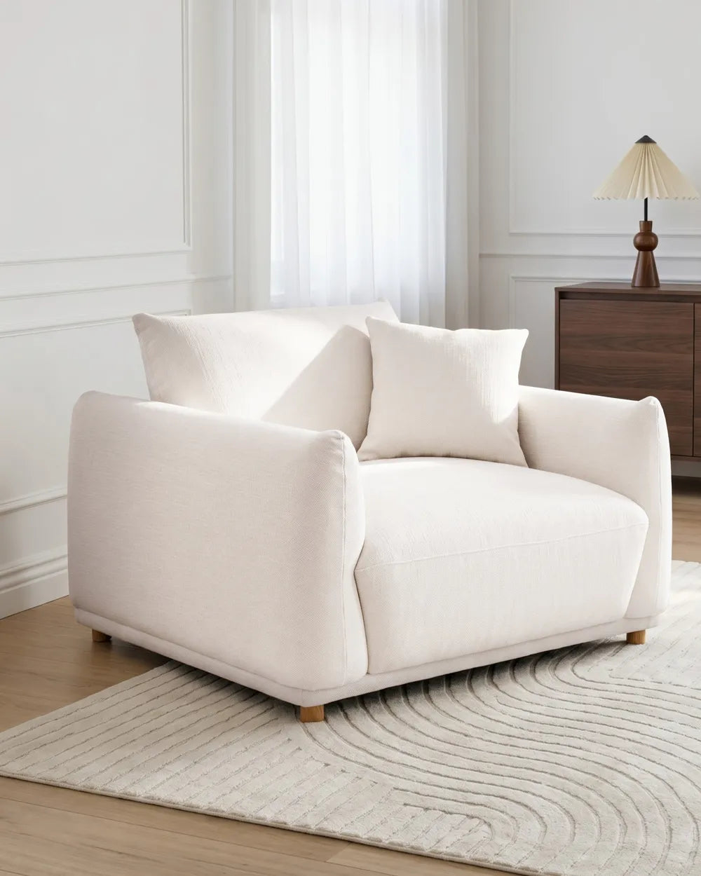 A cream-colored upholstered armchair with two matching throw pillows, set in a bright, modern living room on a textured light rug.