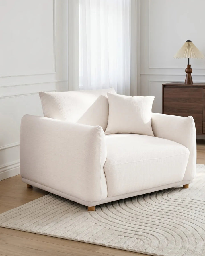 A cream-colored upholstered armchair with two matching throw pillows, set in a bright, modern living room on a textured light rug.