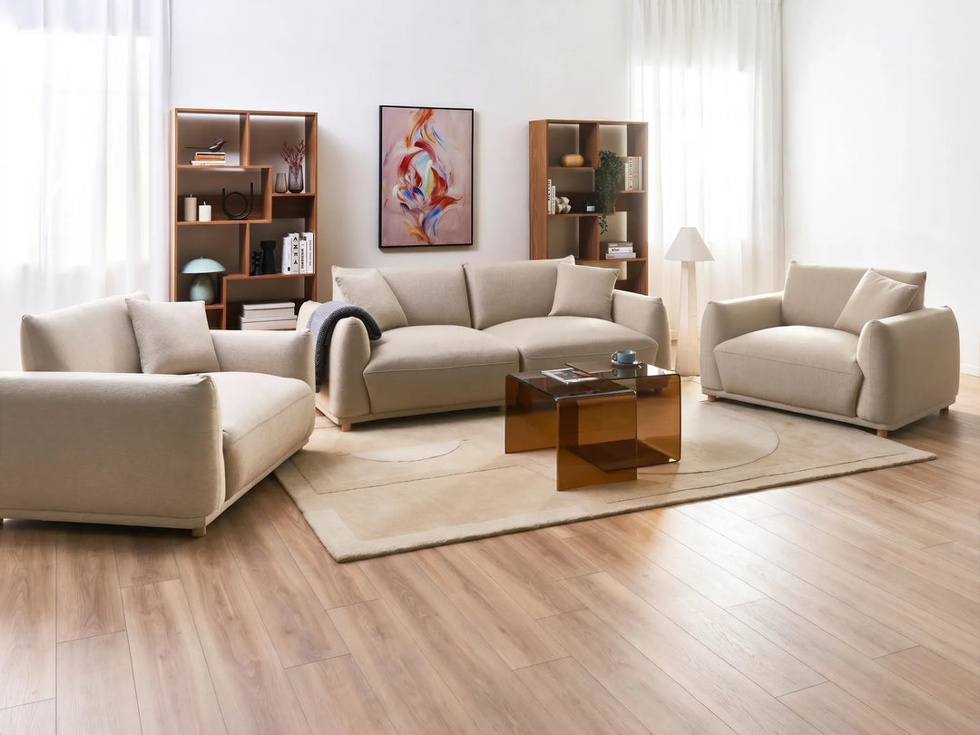 A modern living room set featuring a beige three-seater sofa, a matching armchair, and a chaise lounge arranged around a translucent amber glass coffee table on a light beige rug. The furniture is set on a light wood floor against a white wall with sheer curtains, bookshelves, and a framed abstract print.