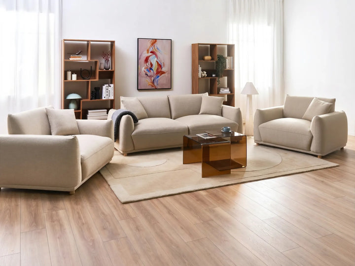 A modern living room set featuring a beige three-seater sofa, a matching armchair, and a chaise lounge arranged around a translucent amber glass coffee table on a light beige rug. The furniture is set on a light wood floor against a white wall with sheer curtains, bookshelves, and a framed abstract print.