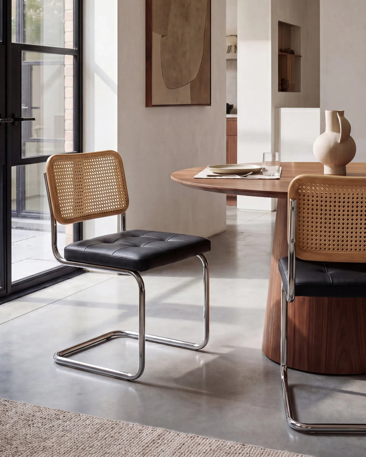 A modern dining room scene featuring two cantilever chairs with black tufted seats and natural woven cane backs, positioned around a round wooden table in a bright, contemporary interior.