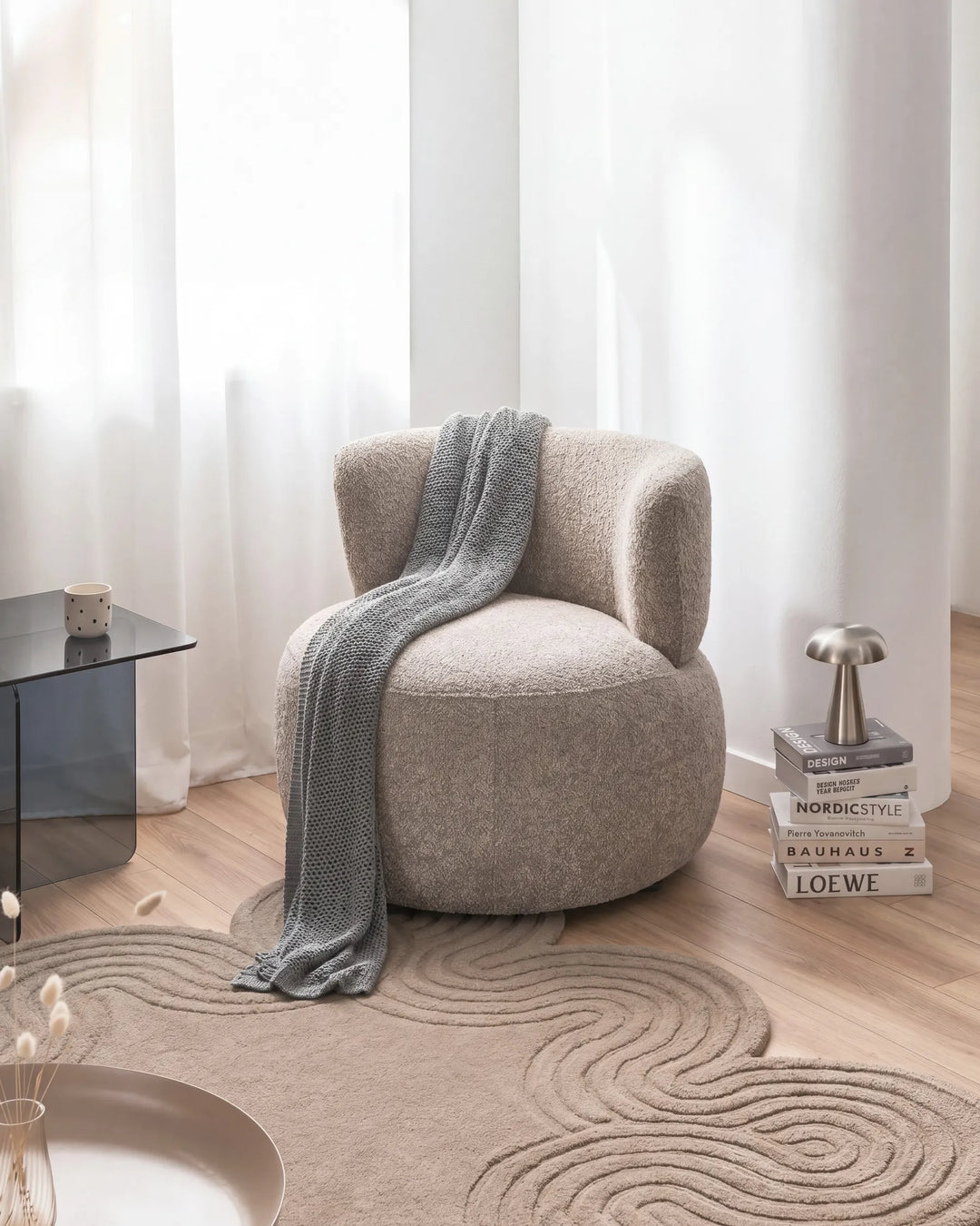 A round upholstered accent chair in a textured light beige fabric, styled with a draped gray knit throw blanket. The chair is set in a bright, modern room with light wood floors, a matching cloud-patterned rug, sheer white curtains, and minimalist decor.