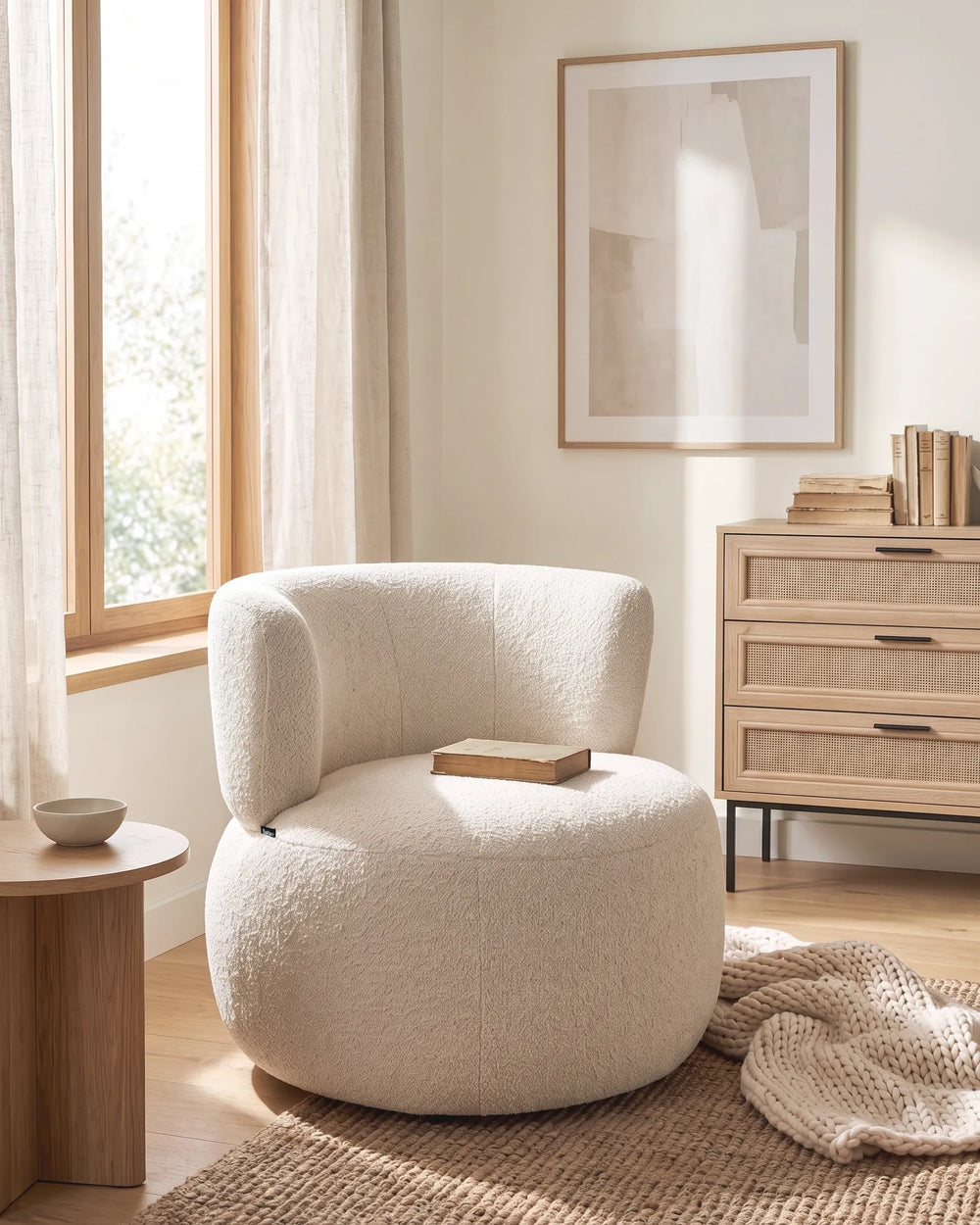 A round, low-profile armchair upholstered in a textured bouclé fabric in a warm off-white or cream color, styled in a bright, modern room next to a wooden side table and a woven rug.