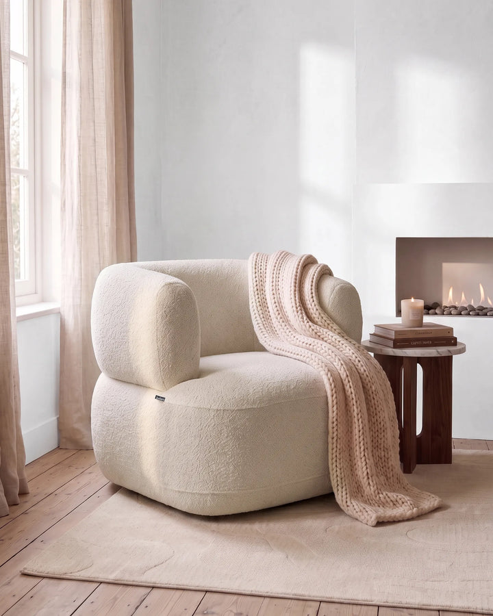 A rounded boucle armchair in warm cream sits in a softly lit room with a beige knit throw draped over it, next to a small wooden side table with books and a candle, and a fireplace is visible in the background.