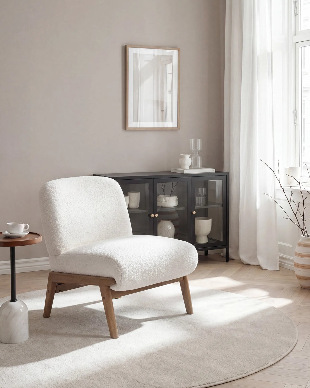 A modern white bouclé accent chair with light wood legs, positioned in a bright, neutral-toned room next to a black glass cabinet and a round white rug.