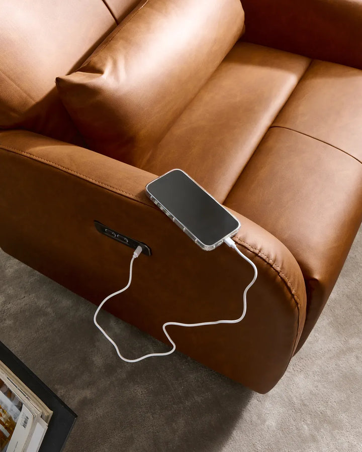 A close-up of a brown leather recliner chair with a side USB charging port, shown with a smartphone plugged in on a gray carpet.