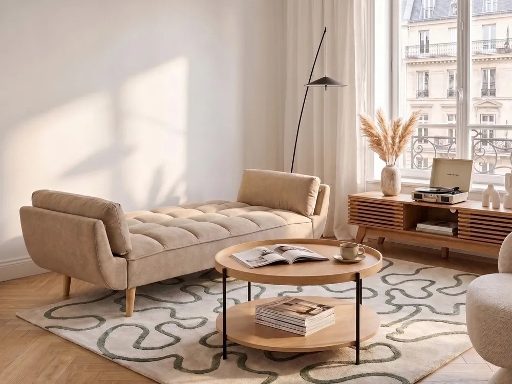 A beige tufted chaise lounge chair in a modern, sunlit living room with a patterned rug, wooden coffee table, and a window view.