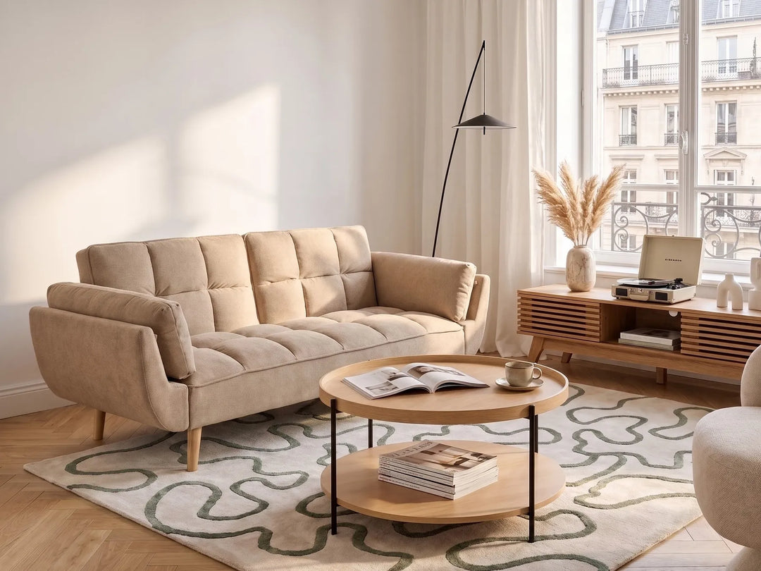 A beige tufted fabric sofa in a bright, modern living room setting with a wooden coffee table, patterned rug, and large window view.