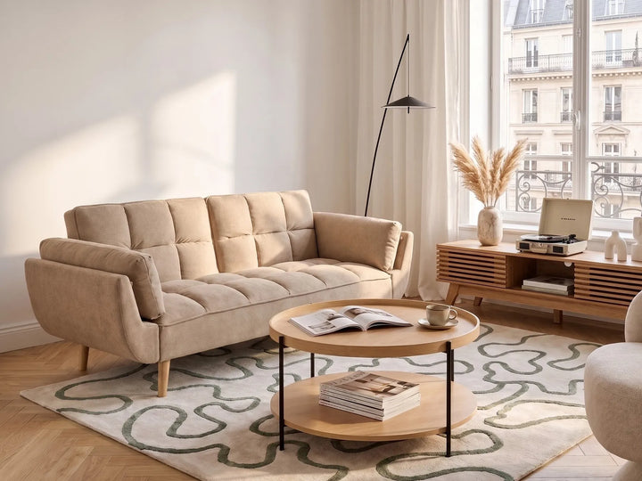 A beige tufted fabric sofa in a bright, modern living room setting with a wooden coffee table, patterned rug, and large window view.
