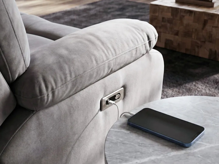 Close-up of a light gray upholstered recliner chair with a side power outlet, shown in a living room setting with a charging smartphone placed on a round table.
