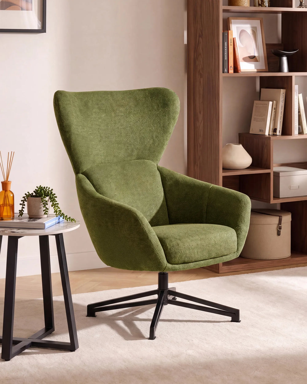 An olive green upholstered swivel armchair with a high curved back and black metal star base, shown in a styled living room setting next to a small side table and a wooden bookshelf.