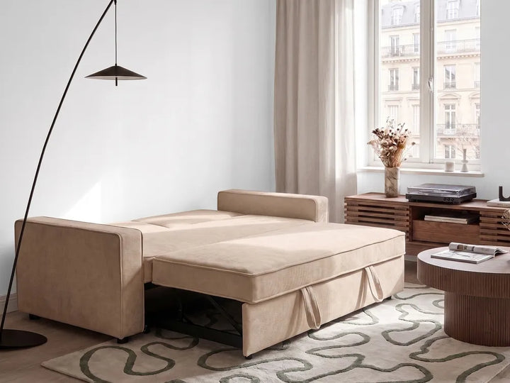 A beige upholstered daybed or sofa bed shown in a styled living room setting, with the seat cushion pulled out to reveal the support frame underneath. The room includes a black floor lamp, a window with curtains, a wooden side table, and a patterned rug.