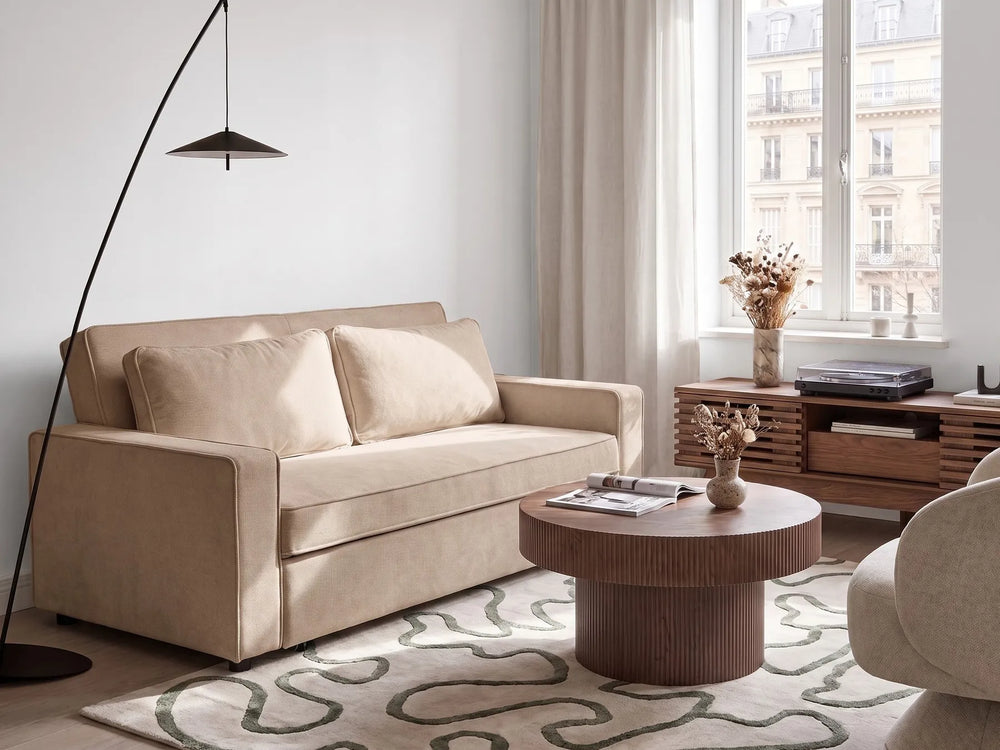 A beige upholstered loveseat sofa in a modern, sunlit living room. The sofa is styled with two matching throw pillows, positioned next to a black floor lamp, a round wooden coffee table, and a patterned rug.