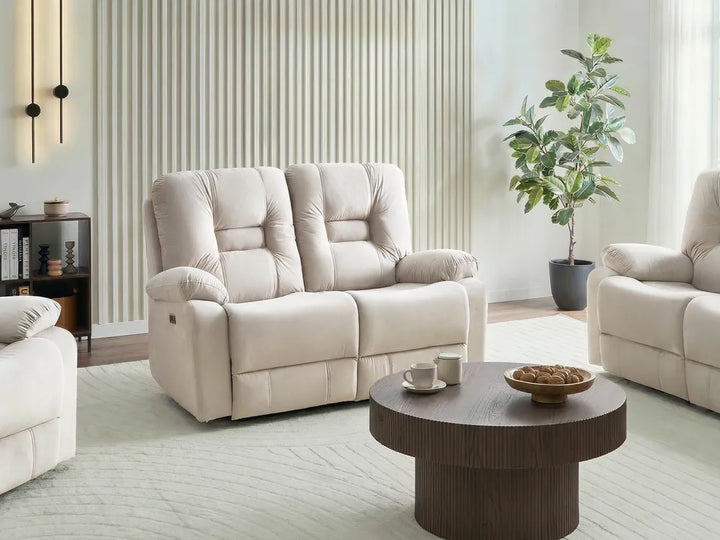 A cream-colored two-seater reclining loveseat sofa in a modern living room setting, styled with a matching armchair, a round wooden coffee table, and a potted plant.