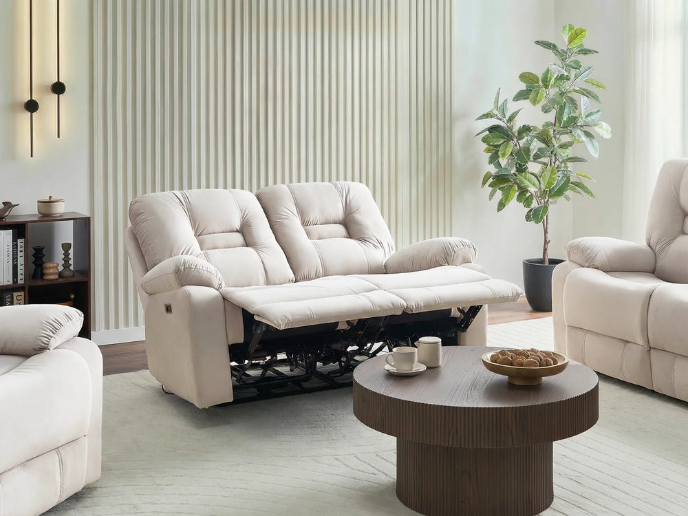 A cream-colored two-seater reclining sofa shown in a modern living room setting with a matching armchair, a round wooden coffee table, and a potted plant.