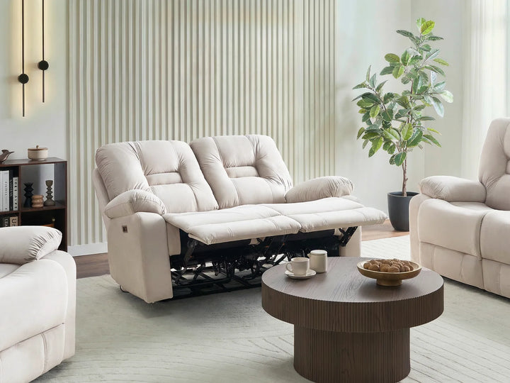 A cream-colored two-seater reclining sofa shown in a modern living room setting with a matching armchair, a round wooden coffee table, and a potted plant.