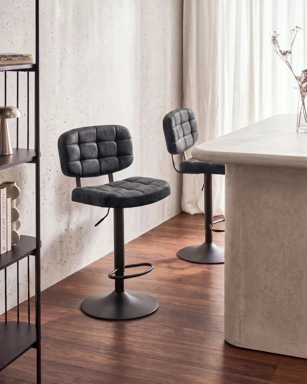 Two black tufted bar stools with adjustable metal bases and footrests, placed in a modern interior with wood flooring, a concrete wall, and a light stone counter.