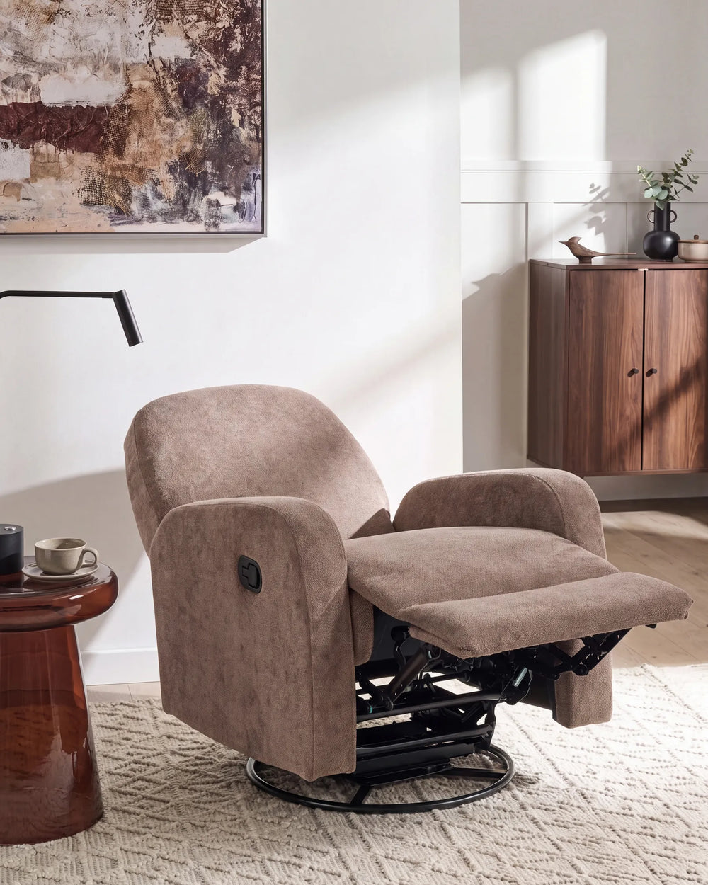 A plush upholstered recliner chair in a warm taupe-brown fabric, shown reclined in a styled living room with a wooden cabinet, wall art, and a rug.