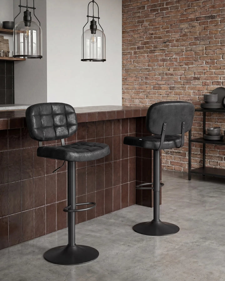 Two black bar stools with tufted seats and metal pedestal bases, placed in a modern kitchen or bar setting with a brown tiled counter, exposed brick wall, and pendant lights.