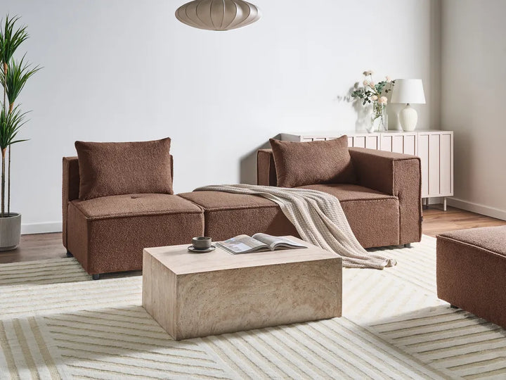 A modern living room setup featuring a brown upholstered modular sofa set with matching cushions, a textured beige stone coffee table, a striped area rug, and a light wood sideboard in the background.