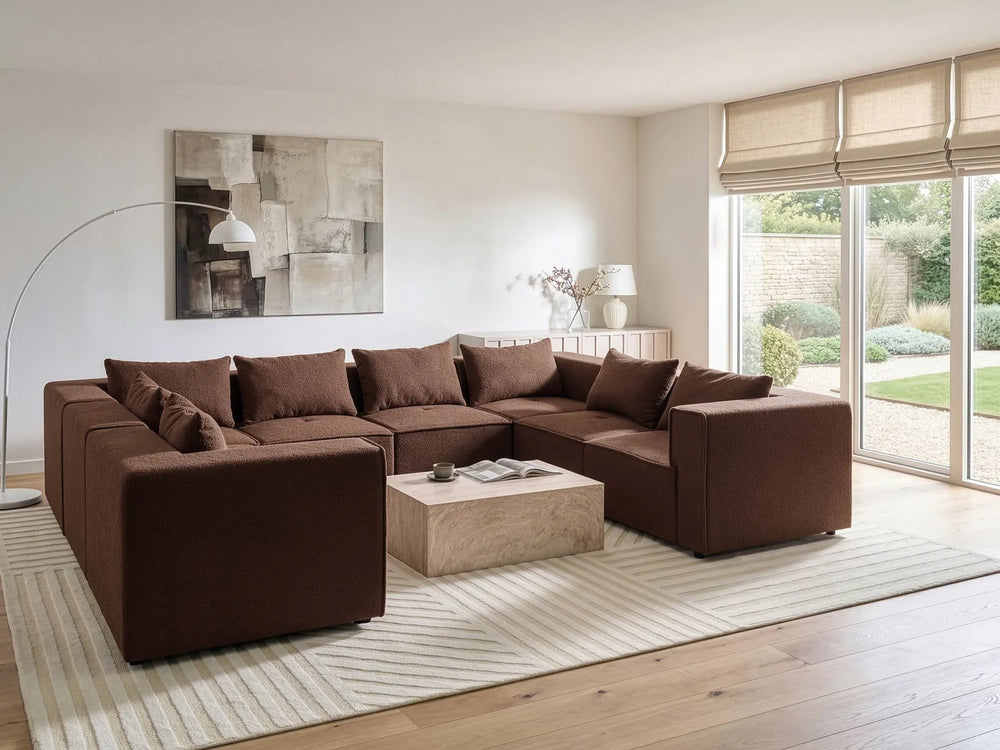 A large L-shaped sectional sofa upholstered in dark brown fabric, styled in a modern living room with a light wood coffee table, striped area rug, and large windows.