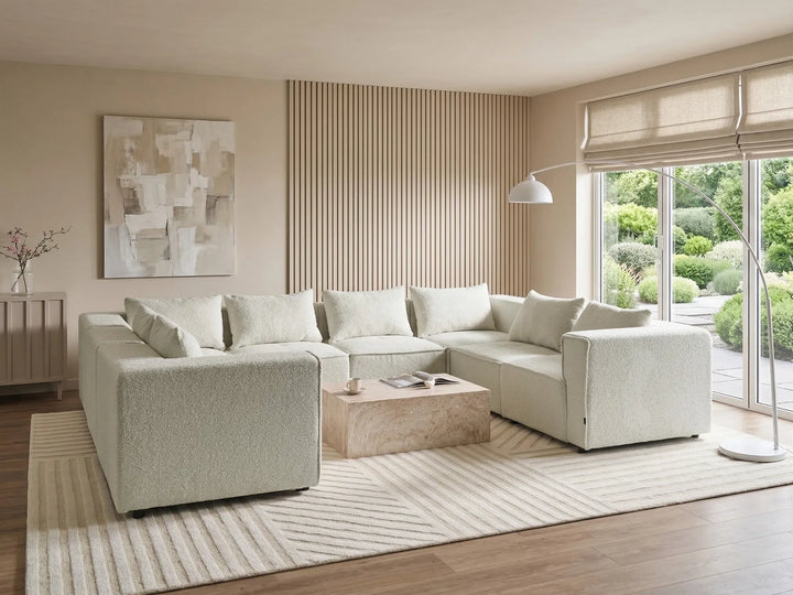 A modern light beige textured sectional sofa with matching throw pillows, styled in a bright, contemporary living room with a striped rug, wooden slat accent wall, and large windows overlooking a garden.