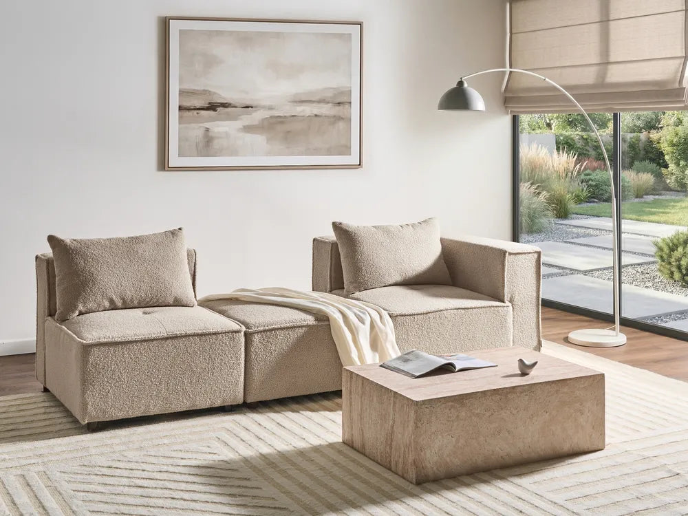 A modern beige sectional sofa with two matching throw pillows and a cream throw blanket, styled in a bright, contemporary living room next to a large window with a floor lamp and a low wooden coffee table.