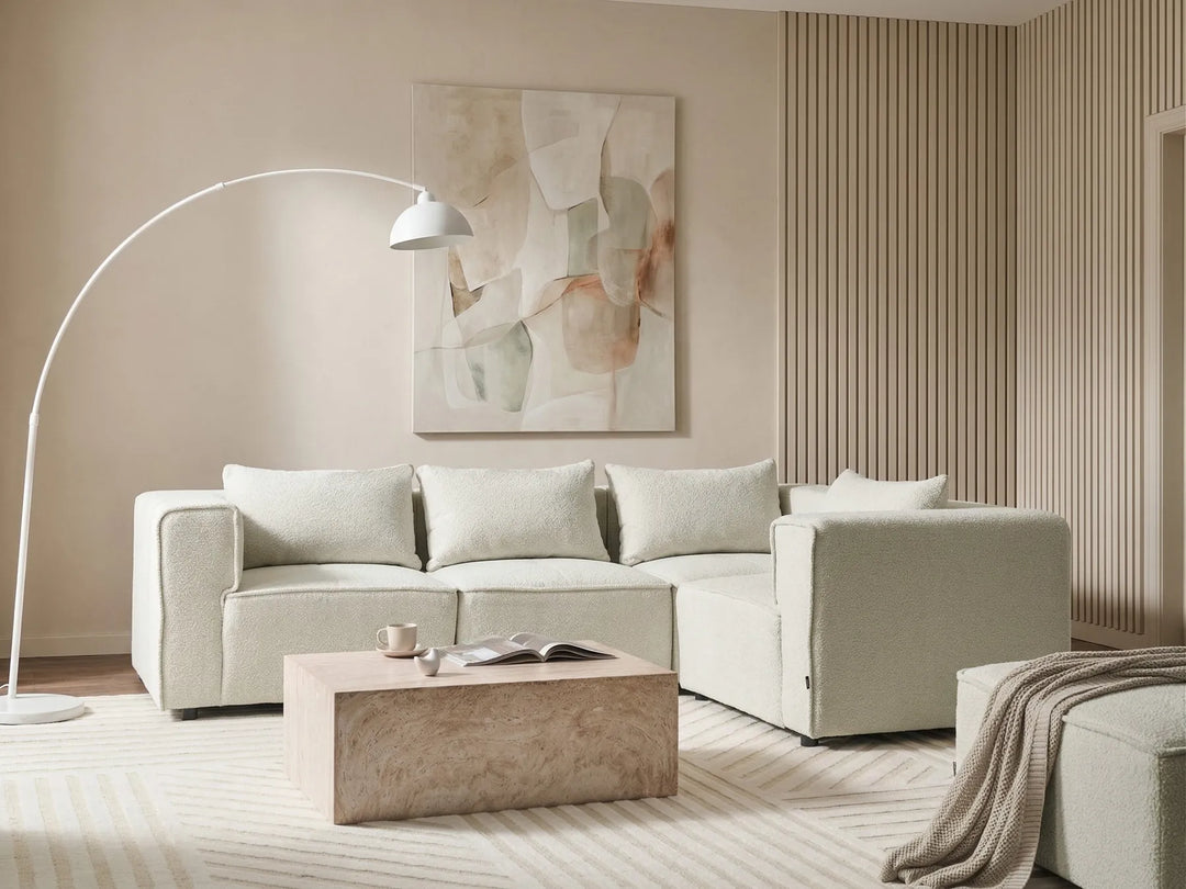 A modern light beige sectional sofa in a minimalist living room setting, styled with a textured stone coffee table, a white floor lamp, and an abstract painting on the wall.