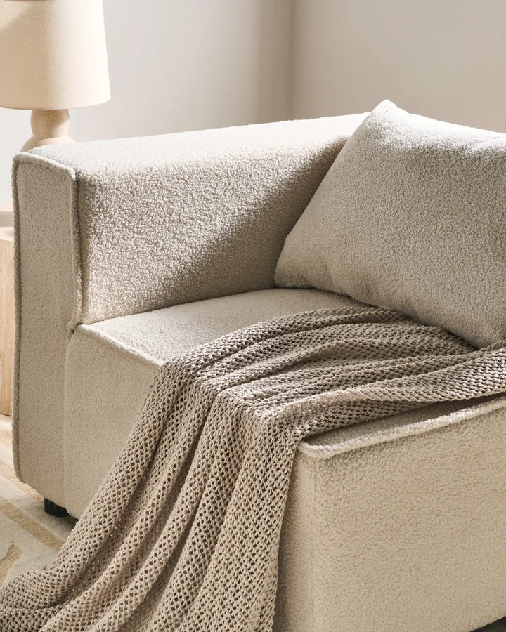 A cozy beige upholstered armchair with a matching textured throw blanket and pillow, styled in a warm, softly lit room with a lamp partially visible in the background.