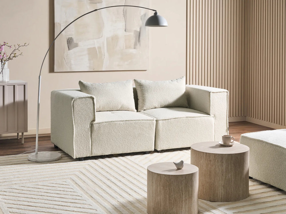 A modern two-seater sofa upholstered in a textured cream bouclé fabric, styled in a minimalist living room with a silver floor lamp, striped rug, and wooden side tables.