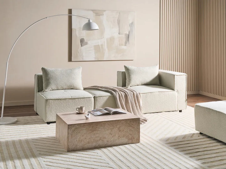 A modern living room setup featuring a light beige sectional sofa with two matching throw pillows, a textured beige coffee table, a tall white floor lamp, and a striped beige and white rug.
