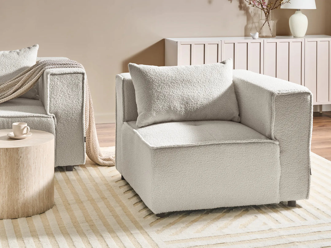 A light beige bouclé armchair with a matching throw pillow, styled in a warm, modern living room setting with a striped rug, wooden side table, and neutral-toned furniture.