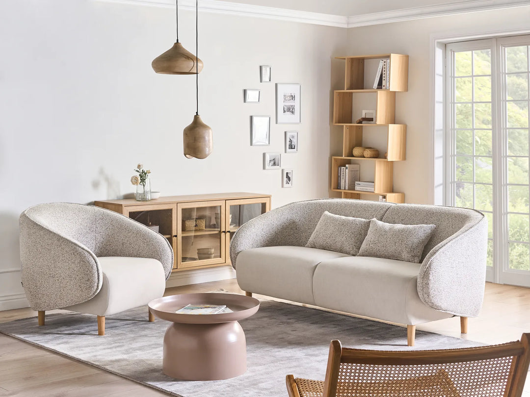 A modern living room featuring a light beige upholstered loveseat with curved arms, a matching armchair, a round pink side table, and a gray rug, set against a neutral wall with a wooden cabinet and shelving.