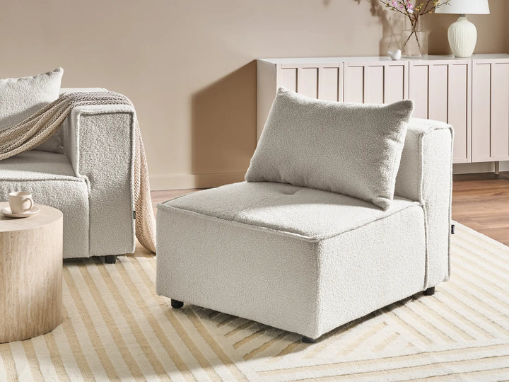 A modern upholstered accent chair in a textured cream bouclé fabric with a matching back cushion, styled in a warm, contemporary living room setting.
