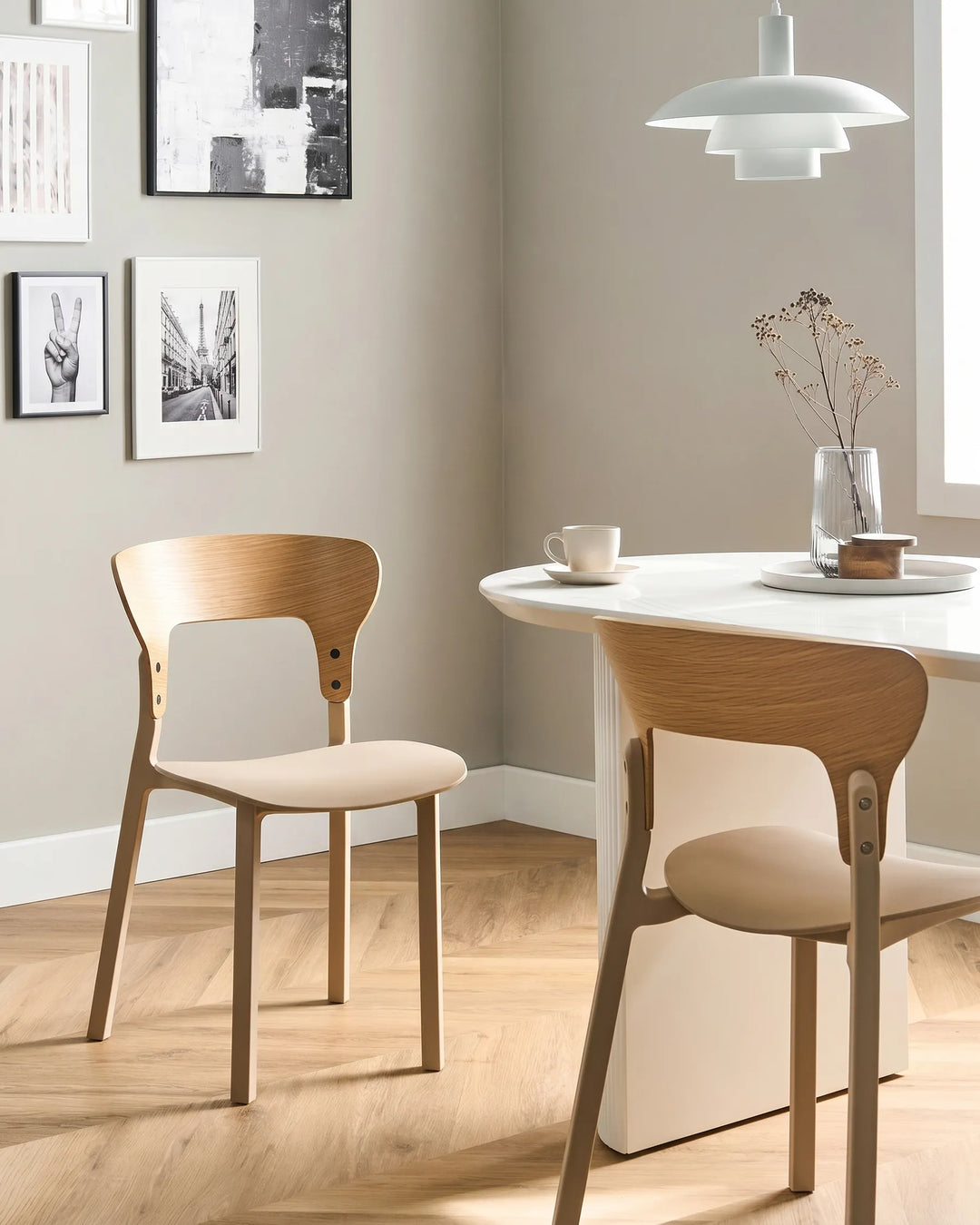 Two modern wooden dining chairs with light beige upholstered seats, shown in a bright, minimalist dining room next to a white table and under a white pendant light.