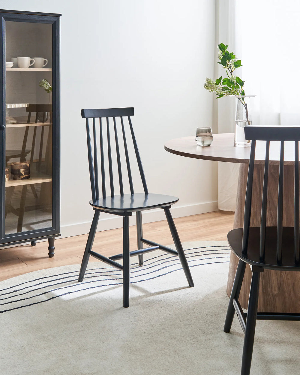 A black spindle-back dining chair is shown in a bright, modern dining room setting next to a round wooden table and another chair, with a tall dark cabinet in the background.