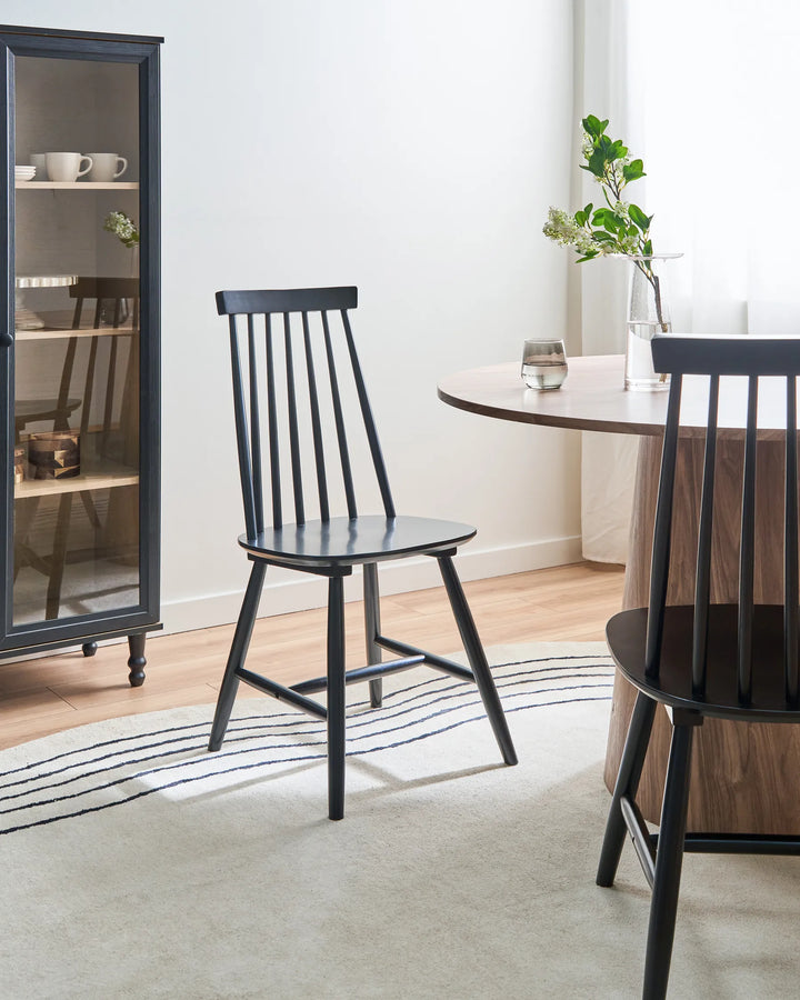 A black spindle-back dining chair is shown in a bright, modern dining room setting next to a round wooden table and another chair, with a tall dark cabinet in the background.