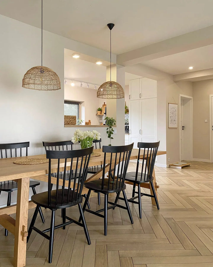 A dining room scene with a light wood dining table and five black spindle-back chairs, under two woven wicker pendant lights. The room has a herringbone wood floor, white walls, and an open kitchen area in the background.