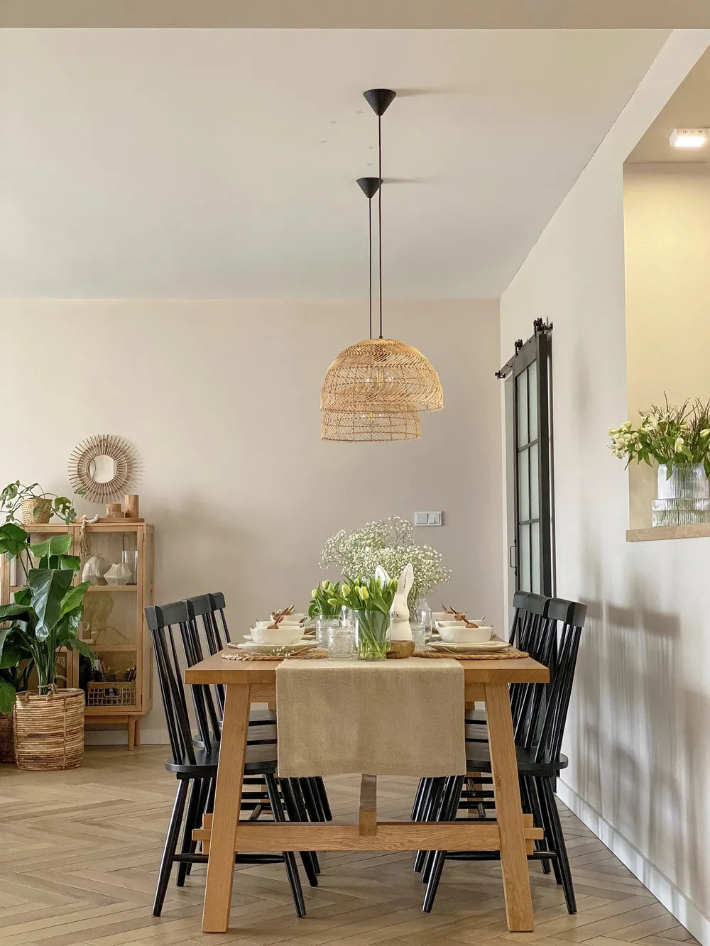A modern dining room featuring a light wood dining table with black spindle-back chairs, a beige table runner, and a woven wicker pendant light hanging above. The room has light beige walls, wood herringbone flooring, and is decorated with plants and a wooden side cabinet.