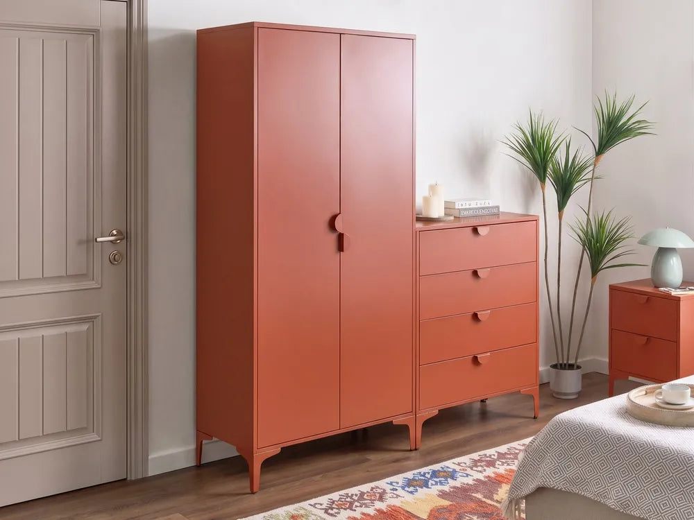 A rust-orange wooden wardrobe and matching five-drawer chest in a styled bedroom setting with a patterned rug, potted plant, and bedside table.
