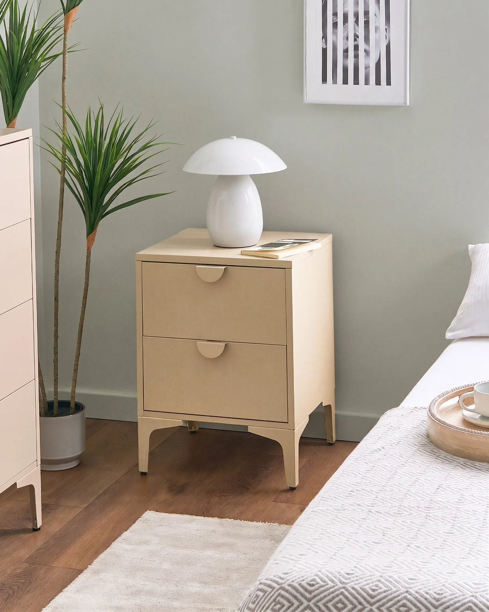 A light beige two-drawer nightstand in a styled bedroom setting, topped with a white mushroom-shaped table lamp, next to a bed and a potted plant.