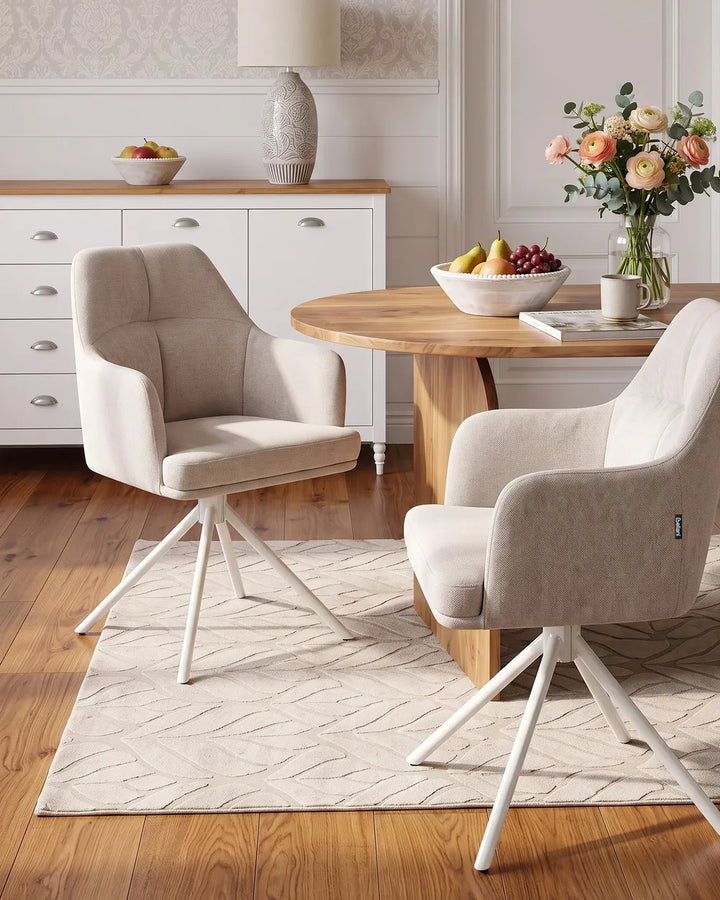 Two modern upholstered dining chairs in a light beige fabric with white swivel legs, arranged around a round wooden table in a bright, contemporary dining room.