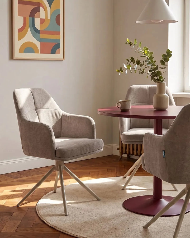 A modern dining room scene featuring two light gray upholstered swivel armchairs with metal star bases, arranged around a round magenta table on a cream rug. A third chair is partially visible, along with a vase of greenery, a mug, and a framed abstract painting on the wall.