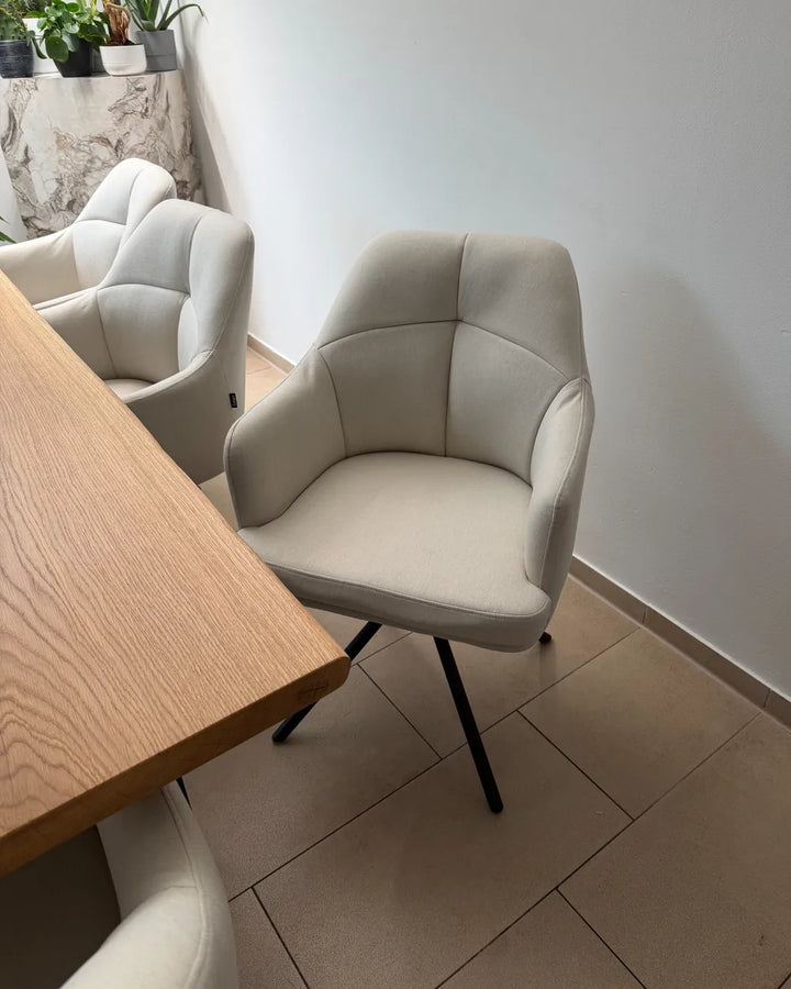 A modern light beige upholstered dining chair with black metal legs, positioned next to a wooden table in a minimalist room with tiled floors and other matching chairs.